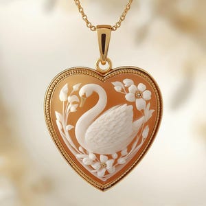 Puede incluir: Collar dorado con colgante en forma de corazón. El colgante presenta un camafeo tallado de un cisne y flores. El cisne es blanco, sobre un fondo naranja claro. El colgante está enmarcado en oro y suspendido de una delicada cadena.