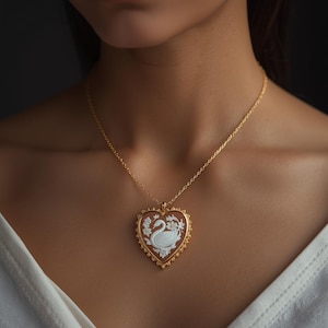 Op de afbeelding: Een goudkleurige ketting met een hartvormige hanger. De hanger heeft een camee-ontwerp met een witte zwaan en bloemendetails op een bruine achtergrond. De ketting wordt om de nek gedragen.