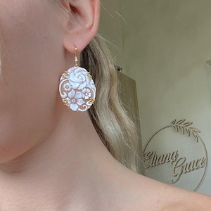 Può includere: Orecchini ovali con un design a cammeo floreale. Gli orecchini presentano un gancio color oro e un'intagliatura dettagliata di fiori e foglie in un colore beige chiaro. Gli orecchini sono indossati all'orecchio di una persona.