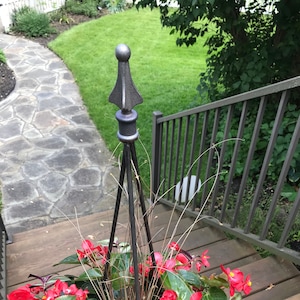 May include: A decorative black metal plant stand with a pointed top, holding vibrant red flowers and green foliage. The stand is on a wooden deck, with a stone pathway and green lawn in the background.