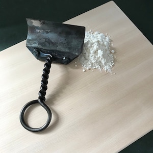 May include: A hand-forged metal dough scraper with a twisted handle and a ring at the end. The scraper is next to a pile of white powder, likely flour, on a light-colored wooden surface.