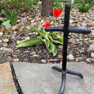 May include: A black metal cross with a textured finish stands outdoors. The cross is positioned in front of red tulips and a backdrop of rocks and greenery. The cross has a simple, yet elegant design.