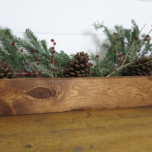 May include: A rustic wooden planter box filled with evergreen branches, pinecones, red berries, and feathers. The box is stained a warm brown and sits on a wooden surface, creating a natural, seasonal centerpiece.