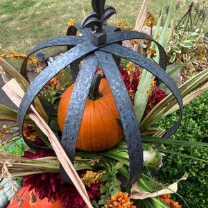 以下が含まれることがあります： 中央にオレンジ色のカボチャ、周囲に秋の葉をあしらった装飾的な金属製の王冠。王冠は濃いグレーで、テクスチャード加工が施されており、上部にはフルール・ド・リスのデザインが施されています。オレンジ、赤、緑のアクセントが含まれています。