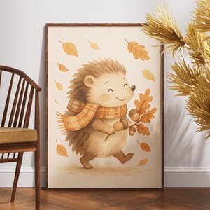Könnte beinhalten: Ein gerahmter Aquarelldruck mit einem fröhlichen Igel, der einen Schal mit Karomuster trägt und Herbstblätter hält. Das Kunstwerk hat eine warme Farbpalette aus Braun- und Orangetönen, mit fallenden Blättern im Hintergrund. Der Druck wird in der Nähe eines Holzstuhls und dekorativer Trockenpflanzen ausgestellt.