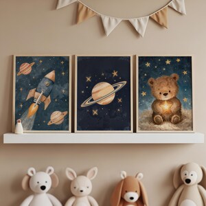 May include: Three framed watercolour art prints with a space theme. The prints depict a rocket, Saturn, and a teddy bear holding a star, set against a dark blue starry background. The frames are light brown.