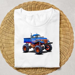 Puede incluir: Camiseta blanca con una ilustraci&oacute;n de dibujos animados de un cami&oacute;n monstruo azul con llamas naranjas y amarillas. El cami&oacute;n tiene neum&aacute;ticos negros grandes con llantas naranjas. La camiseta se muestra sobre una superficie circular tejida.