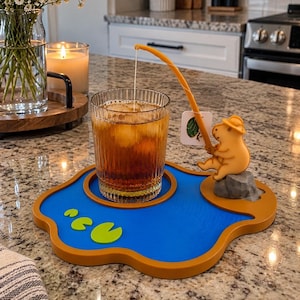 May include: A blue and brown pond-shaped coaster featuring a capybara figurine sitting on a gray rock. The capybara holds a yellow fishing rod that suspends a tea bag tag over a glass of iced tea. Two small green lily pads are on the blue surface.