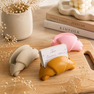 May include: Three croissant-shaped card holders in beige, mustard yellow, and light pink. The mustard yellow holder contains a white rectangular business card with the handwritten text "Your Business card" and small heart and croissant illustrations.