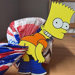 May include: A flat, yellow cartoon character cutout featuring a spiky-haired boy wearing an orange shirt and blue pants pulled down, positioned in front of a plastic bread bag with red, white, and blue patterns and printed text.