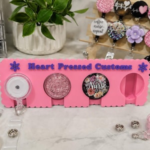 May include: A pink rectangular display stand with scalloped edges featuring the text Heart Pressed Customs in purple. The stand has circular cutouts holding badge reels, including one clear reel, one pink glitter reel, and one floral nurse reel.