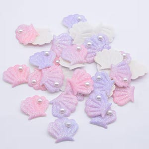 Könnte beinhalten: Eine Sammlung pastellfarbener Muschelschmuckstücke mit Glitzer- und Perlenakzenten. Die Muscheln sind in Rosa- und Lavendeltönen gehalten und haben eine weiße Stoffrückseite. Ideal für Bastelprojekte.
