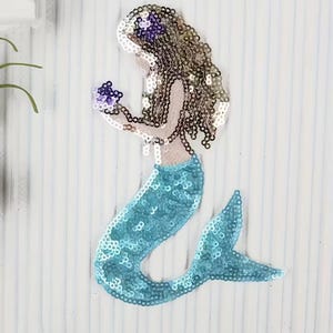 Könnte beinhalten: Ein meerjungfrauenförmiger Aufnäher mit Paillettenbesatz. Die Figur hat langes, gewelltes braunes Haar mit einer violetten Blume, einen hellbeigen Oberkörper und einen leuchtend blauen Paillettenschwanz. Die Meerjungfrau hält eine kleine violette Blume.
