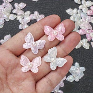 Könnte beinhalten: Nahaufnahme von mehreren schmetterlingsförmigen Verzierungen. Die Schmetterlinge sind in Rosa- und Weißtönen gehalten und haben jeweils eine Perle in der Mitte. Einige werden in einer Hand gehalten, andere sind auf einer Oberfläche verstreut.