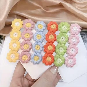 Könnte beinhalten: Sechs Haarspangen mit vertikalen Reihen gestickter Blumen in Gelb, Rosa, Hellblau, Orange, Limettengrün und zartem Rosa. Jede Blume hat eine kontrastierende Mitte, und die Spangen sind nebeneinander auf einer weißen Karte angeordnet.
