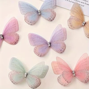 Könnte beinhalten: Eine Sammlung von sechs Schmetterlingsornamenten aus transparentem Stoff in Pastelltönen wie Blau, Gelb, Rosa, Lila und Grün. Jeder Schmetterling hat geschichtete Flügel mit einem dezenten Punktmuster und einen mit kleinen klaren Kristallen besetzten Körper.