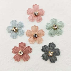 Könnte beinhalten: Sechs Stoffblumen-Verzierungen in verschiedenen Pastellfarben: Rosa, Pfirsich, Blau, Grün und Grau. Jede Blume hat einen funkelnden Strassstein in der Mitte. Diese dekorativen Blumen eignen sich ideal zum Basteln und verleihen Projekten Eleganz.