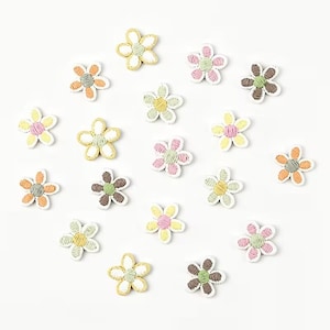 Könnte beinhalten: Eine Ansammlung kleiner, fünfblättriger blumenförmiger Verzierungen mit gestickten Faden-Details in verschiedenen Pastellfarben wie Rosa, Gelb, Grün, Orange und Braun, die verstreut auf einem schlichten weißen Hintergrund angeordnet sind.