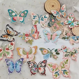 Könnte beinhalten: Eine Sammlung von fünfzehn bestickten Schmetterlingsaufnähern in verschiedenen Farbpaletten, darunter Rosa, Blau, Grün, Orange und Weiß. Jeder Aufnäher zeigt komplizierte Blumen- und Blattmuster, die auf die Flügel der Schmetterlinge gestickt sind.