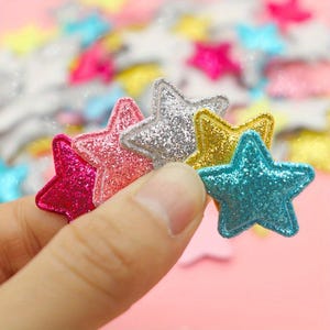 Könnte beinhalten: Eine Sammlung kleiner, fünfzackiger sternförmiger Aufnäher mit glitzernder Oberfläche. Die Sterne sind in verschiedenen leuchtenden Farben erhältlich, darunter Pink, Hellrosa, Silber, Gold und Türkis, jeweils mit einem genähten Rand.