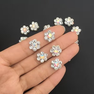 Könnte beinhalten: Kleine, blütenförmige Verzierungen mit schillernden Zentren und klaren, funkelnden Rändern. Diese dekorativen Teile sind auf einem dunklen Hintergrund verstreut. Die floralen Akzente sind ideal für Bastelarbeiten und DIY-Projekte.
