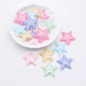 Könnte beinhalten: Eine Sammlung kleiner, fünfzackiger sternförmiger Applikationen in verschiedenen Pastellfarben wie Rosa, Blau, Gelb, Lila und Grün. Jeder Stern hat eine strukturierte, glitzernde Oberfläche und ein erhabenes, gepolstertes Aussehen mit genähten Kanten.