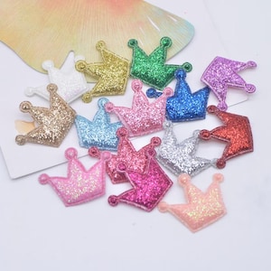 Könnte beinhalten: Eine Sammlung kleiner kronenförmiger Aufnäher, die mit Glitzer bedeckt sind. Die Kronen erscheinen in verschiedenen Farben, darunter Gold, Silber, Grün, Blau, Rot, Pink, Lila und Pfirsich. Jede Krone hat ein dreizackiges Design mit kleinen Akzenten.
