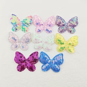 Könnte beinhalten: Eine Sammlung von acht schmetterlingsförmigen Aufnähern, die mit bunten Pailletten verziert sind. Das Set enthält Schmetterlinge in irisierendem Weiß, Hellrosa, Lila, Gelb, Magenta und Blau, jeweils mit einer Netzbasis, die mit glänzenden Pailletten bedeckt ist.