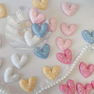 Könnte beinhalten: Eine Sammlung glitzernder herzförmiger Verzierungen in Pink, Blau, Gelb und Weiß, verstreut neben einer transparenten, muschelförmigen Kunststoffschale und einem weißen Spitzenband mit kleinen Perlen. Die Schale zeigt einen blassen Goldtext.