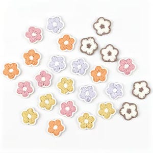 Könnte beinhalten: Eine Sammlung kleiner, fünfblättriger Blumenaufnäher mit detaillierter Stickerei. Das Set enthält verschiedene Farben wie zartes Rosa, helles Orange, blasses Gelb, Lavendel und Weiß, jeweils mit einem kontrastierenden Detail in der Mitte.