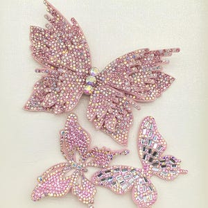 Könnte beinhalten: Drei schmetterlingsförmige Applikationen in verschiedenen Größen, gefertigt aus hellrosa Material und dicht besetzt mit schimmernden rosa und irisierenden Strasssteinen. Der größte Schmetterling hat drei markante runde Kristalle in der Mitte.
