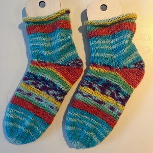 Könnte beinhalten: Ein Paar handgestrickte Socken mit einem lebendigen, mehrfarbigen Design. Die Socken haben eine blaue Basis mit horizontalen Streifen in Rot, Gelb, Grün und Lila. Die Bündchen sind gelb, und die Fersen und Zehen sind rot. Jede Socke ist an einem kleinen Holzanhänger befestigt.