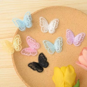 Könnte beinhalten: Eine Sammlung von sieben bestickten Schmetterlingsaufnähern in verschiedenen Farben, darunter Hellblau, Weiß, Gelb, Rosa und Schwarz. Jeder Aufnäher weist eine detaillierte Fadenstickerei auf den Flügeln und eine feine Netzstruktur auf.