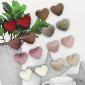 Könnte beinhalten: Eine Sammlung kleiner, plüschiger herzförmiger Ornamente in verschiedenen Rot-, Braun-, Beige- und Rosatönen. Einige Herzen sind an grünen Blattstielen in einem hohen Metallbehälter befestigt, andere sind vor einem weißen Hintergrund angeordnet.