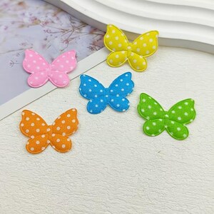 Könnte beinhalten: Fünf Schmetterlings-Applikationen aus Stoff in Gelb, Pink, Blau, Orange und Grün, jeweils mit einem weißen Punktmuster versehen. Diese kleinen, flachen, textilen Schmetterlingsformen sind in einer verstreuten Anordnung platziert.