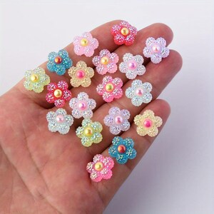 Könnte beinhalten: Eine Sammlung kleiner, fünfblättriger blumenförmiger Verzierungen mit einer strukturierten, glitzernden Oberfläche und einer glatten, runden, perlenartigen Mitte. Diese dekorativen Teile sind in verschiedenen Pastell- und leuchtenden Farben wie Pink, Blau, Gelb und Weiß erhältlich.