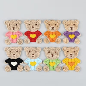 Könnte beinhalten: Acht gehäkelte Bären-Applikationen in zwei Reihen angeordnet. Jeder Bär ist hellbraun mit dunkelbraun gestickten Augen und Nasen. Sie tragen Pullover in Pink, Rot, Orange, Lila, Schwarz, Hellblau, Hellgrün und Gelb mit Herzmotiven.