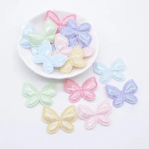 Könnte beinhalten: Eine Sammlung glitzernder schmetterlingsförmiger Applikationen in Pastelltönen wie Rosa, Blau, Gelb, Grün und Lila. Mehrere Schmetterlinge sind in einer weißen, gewellten Schale angeordnet, während weitere um die Basis herum verstreut sind.