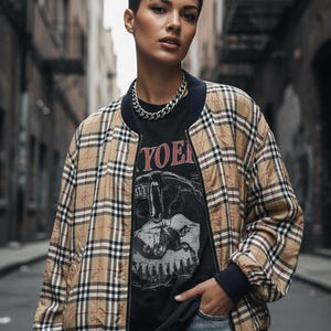 May include: A tan and black plaid bomber jacket with navy blue trim is worn over a black graphic tee. A silver chain necklace is visible. The person is wearing blue jeans.