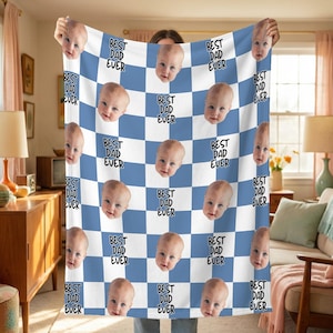 May include: A personalized blanket featuring a repeating pattern of a baby's face and the text "BEST DAD EVER" on a blue and white checkered background. The blanket is held up, showcasing the design.