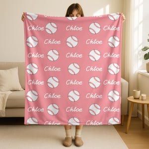 May include: A pink blanket with a repeating pattern of white baseballs with red stitching and the name "Chloe" in white script. The blanket is being held up by a person. The room has a beige sofa and wooden side table.