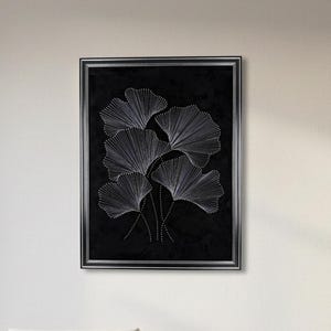 May include: A framed artwork with a black backdrop, showcasing a design of white, fan-shaped leaves. The leaves are crafted with fine white lines, and the frame is silver. The artwork is displayed on a white wall above a white sofa.