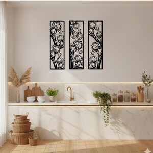 May include: Three black metal wall art panels, each featuring a floral iris design. The panels are rectangular and mounted on a white wall above a kitchen counter. The kitchen has a marble countertop, a gold faucet, and various decorative items.
