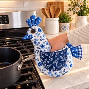 May include: A chicken-shaped oven mitt made of white fabric with a blue floral pattern. It features a dark blue wing-shaped pocket, a blue and white checkered tail, a dark brown button eye, and a blue floral crest on top of the head.