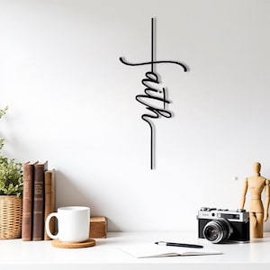 May include: Black metal wall art in the shape of a cross with the word "faith" in a cursive font. The artwork is mounted on a white wall. A white mug, books, and a camera are on a white desk.