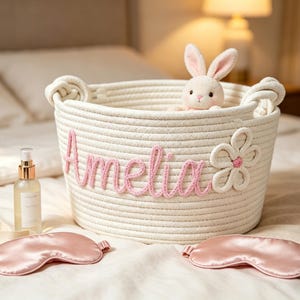 May include: A cream-colored rope basket with the name "Amelia" in pink script, a flower accent, and a stuffed bunny. Two pink satin sleep masks and a bottle of spray are on the bed. The basket has two rope handles.