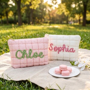 Op de afbeelding: Twee gewatteerde make-up tassen, een roze met "Chloe" in groene letters en een witte met "Sophia" in roze letters. Gouden ritsen en een bord met roze macarons zijn ook zichtbaar.
