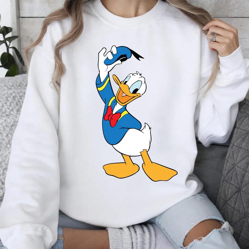May include: White sweatshirt featuring a cartoon illustration of Donald Duck. Donald Duck is depicted in his signature blue sailor suit with a red bow tie, holding his hat. The sweatshirt is a casual style.