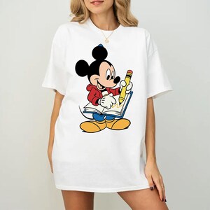 Op de afbeelding: Wit T-shirt met een cartoon illustratie van Mickey Mouse die met een gele potlood in een boek schrijft. Mickey Mouse draagt een rode jas, een blauwe broek en gele schoenen. Het ontwerp is gecentreerd op de voorkant van het shirt.