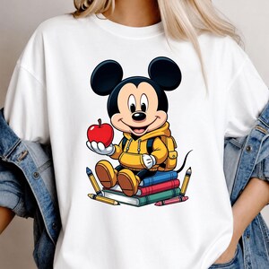 Op de afbeelding: Wit T-shirt met een cartoonontwerp van Mickey Mouse. Mickey draagt een gele hoodie en rugzak, houdt een rode appel vast en zit op een stapel boeken. Potloden en kleurpotloden zijn ook zichtbaar.